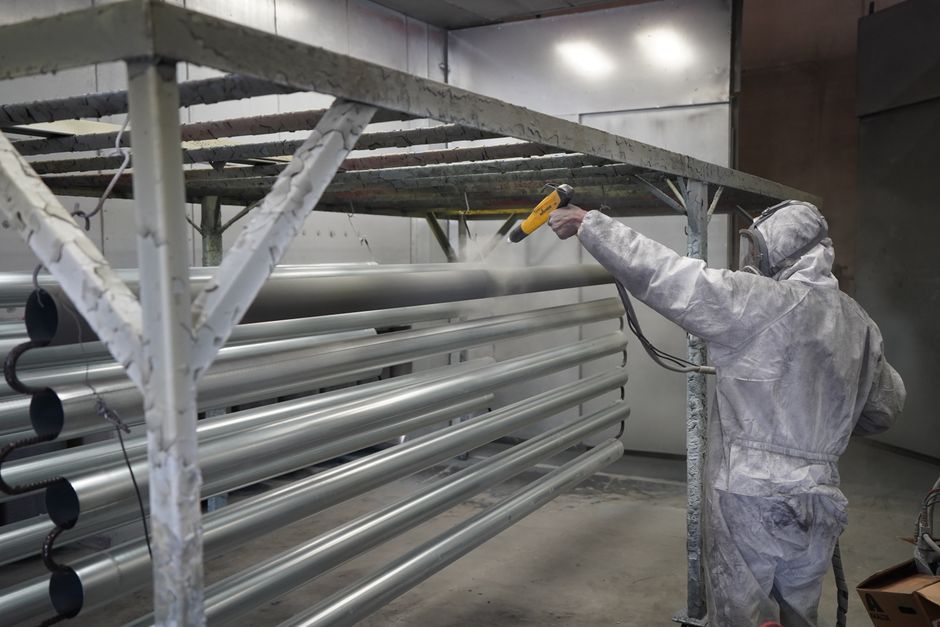 Worker in protective gear applying powder coating to metal pipes in a factory setting.